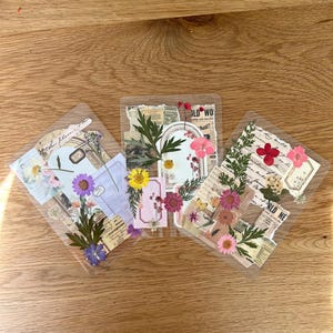 May include: Three clear, rectangular stickers with pressed flowers, paper ephemera, and text. The stickers feature various colors of flowers, including pink, yellow, and purple. The text includes the words "OLD WO" and "hat?"