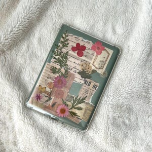 May include: Clear phone case with a collage design featuring pressed flowers, newspaper clippings, and botanical elements. The case has a green background and a variety of pink and yellow flowers. The text "WORLD NEWS" is visible.