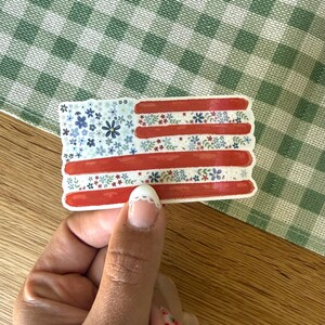Op de afbeelding: Een rechthoekige sticker in de vorm van de Amerikaanse vlag, met een blauw veld vol kleine blauwe en witte bloemen, en horizontale strepen die afwisselen tussen effen rode banen en witte banen versierd met kleine blauwe, witte en groene bloemen.