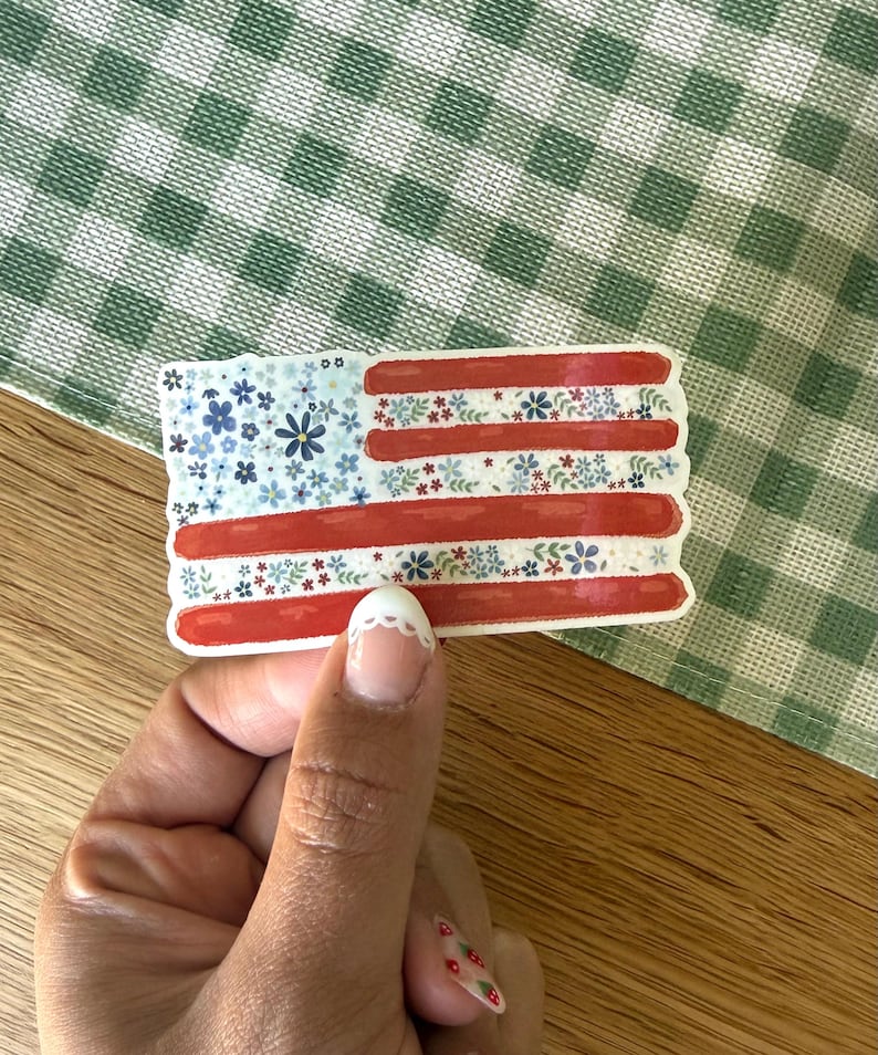 Op de afbeelding: Een rechthoekige sticker in de vorm van de Amerikaanse vlag, met een blauw veld vol kleine blauwe en witte bloemen, en horizontale strepen die afwisselen tussen effen rode banen en witte banen versierd met kleine blauwe, witte en groene bloemen.