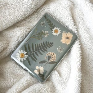 May include: Clear case for a tablet, adorned with pressed flowers and greenery. The design includes white daisies, ferns, and other delicate floral elements, set against a pale green background. The case is rectangular and appears to be made of a transparent material.