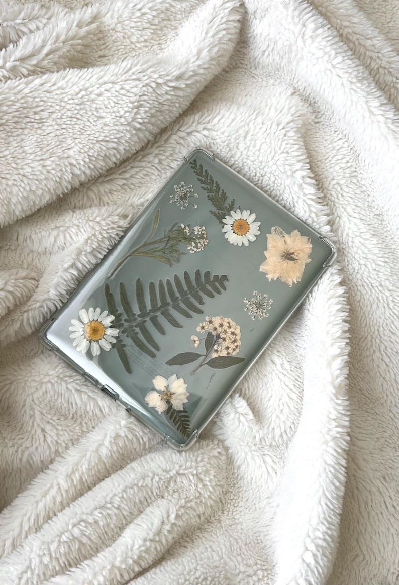May include: Clear case for a tablet, adorned with pressed flowers and greenery. The design includes white daisies, ferns, and other delicate floral elements, set against a pale green background. The case is rectangular and appears to be made of a transparent material.