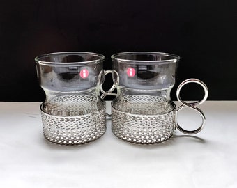 Puede incluir: Dos tazas de vidrio transparente con bandas y asas de metal plateado. Las tazas tienen una pequeña pegatina roja con una letra "I" en ellas.