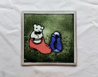 May include: A ceramic tile with a green background featuring a white bear wearing a black bow tie sitting in a red shoe. A blue boot is next to the red shoe.