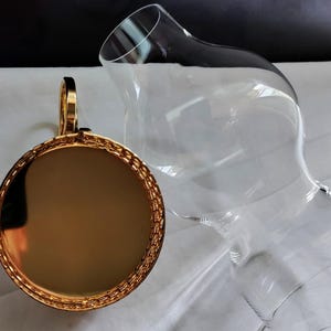 Puede incluir: Un decantador de vidrio transparente con un cuerpo ancho y redondeado y un cuello estrecho, junto con un tap&oacute;n circular de tono dorado que presenta una superficie met&aacute;lica reflectante y un borde texturizado estilo cadena.