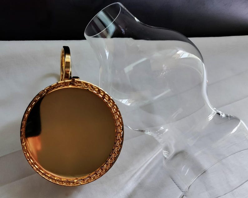 Puede incluir: Un decantador de vidrio transparente con un cuerpo ancho y redondeado y un cuello estrecho, junto con un tap&oacute;n circular de tono dorado que presenta una superficie met&aacute;lica reflectante y un borde texturizado estilo cadena.