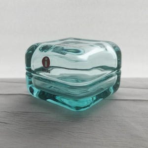 May include: A square, light blue glass box with rounded corners and a thick, clear glass lid. The side of the glass features a small, circular red and white logo with the text iittala printed in black, indicating the brand of this glass container.
