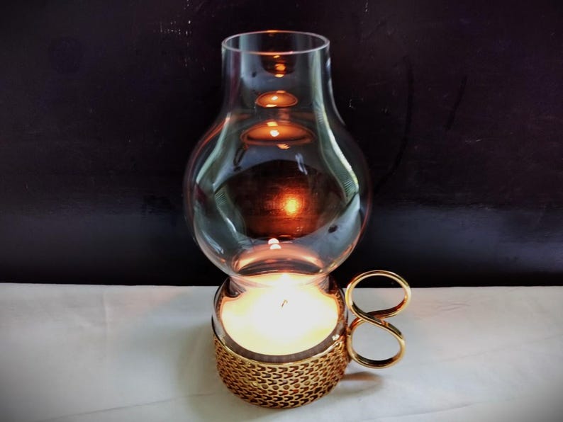 Puede incluir: Un portavelas de cristal con una chimenea transparente y abombada, y una base dorada con un patr&oacute;n texturizado de escamas. La base incluye un asa dorada decorativa en forma de bucle en el lateral, y se ve una vela encendida en el interior.