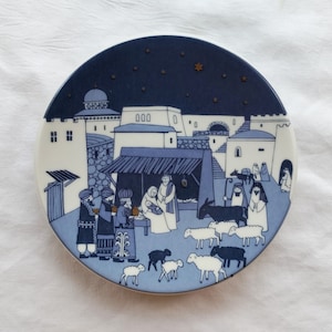 May include: A blue and white ceramic plate with a depiction of the nativity scene. The plate features a blue background with white stars and a white silhouette of a town with a stable in the center. The stable is surrounded by figures of the three wise men, Mary, Joseph, and the baby Jesus. There are also animals, including a donkey and sheep.