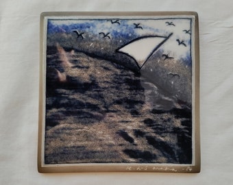 May include: A ceramic tile with a blue and white image of a sailboat on a wavy sea with a flock of birds flying overhead. The tile is signed "E.H.S. Machin - 84".