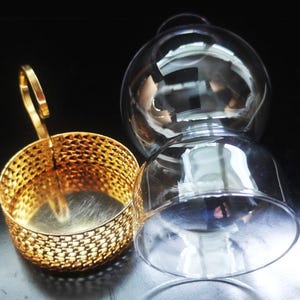 Puede incluir: Un soporte de metal dorado con un patr&oacute;n de malla fina y un asa vertical, colocado junto a un globo de cristal transparente de forma esf&eacute;rica con una amplia abertura en la parte inferior. Ambos componentes son piezas separadas.