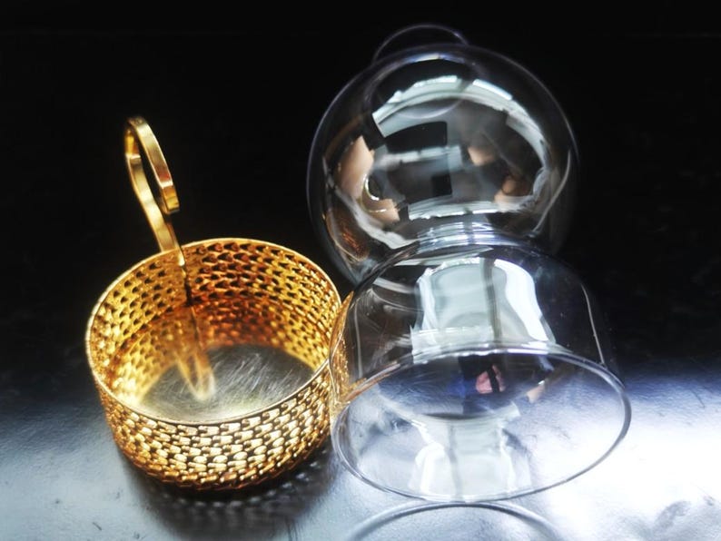 Puede incluir: Un soporte de metal dorado con un patr&oacute;n de malla fina y un asa vertical, colocado junto a un globo de cristal transparente de forma esf&eacute;rica con una amplia abertura en la parte inferior. Ambos componentes son piezas separadas.