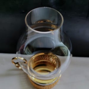 Puede incluir: Una pantalla de l&aacute;mpara de hurac&aacute;n de cristal transparente con un cuerpo redondeado y abombado y una abertura superior ensanchada. La base de la l&aacute;mpara presenta un anillo de metal dorado pulido con un patr&oacute;n texturizado y una peque&ntilde;a asa.