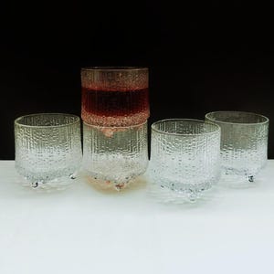 May include: A set of four clear glass tumblers with a textured, bark-like exterior pattern. Each tumbler features three small, rounded feet at the base. One tumbler is stacked on top of another, and the top glass contains a reddish liquid.