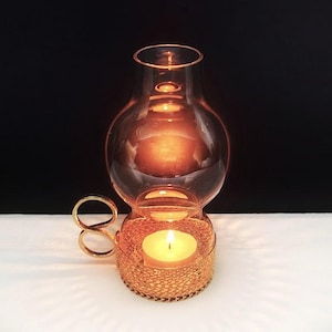 Puede incluir: Un portavelas de metal dorado con una base texturizada tipo malla y un asa en forma de bucle. Una chimenea de vidrio transparente y redondeada se asienta sobre la base, encerrando una vela de t&eacute; encendida que emite un brillo c&aacute;lido y suave.