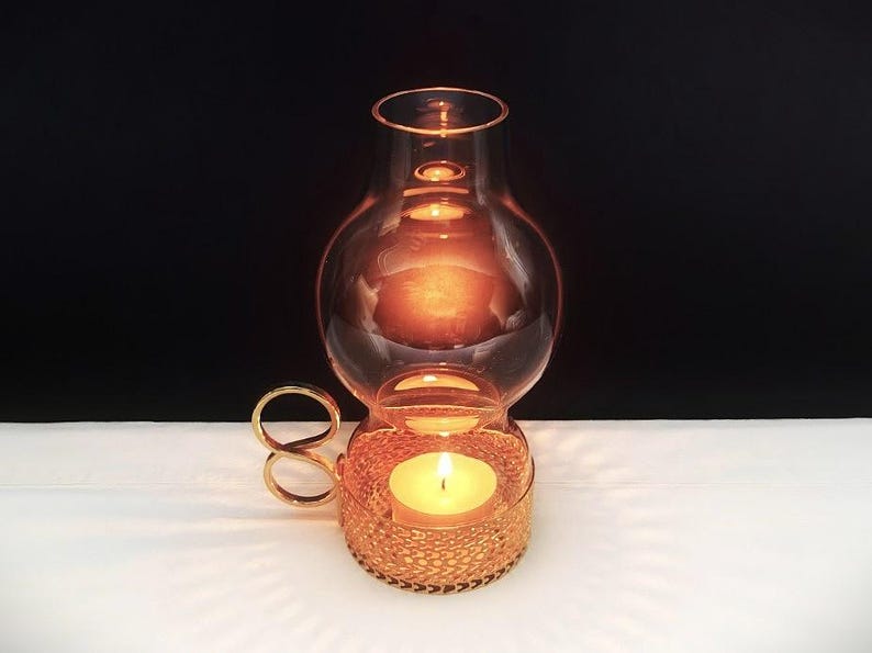 Puede incluir: Un portavelas de metal dorado con una base texturizada tipo malla y un asa en forma de bucle. Una chimenea de vidrio transparente y redondeada se asienta sobre la base, encerrando una vela de t&eacute; encendida que emite un brillo c&aacute;lido y suave.
