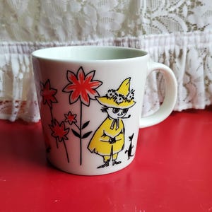 May include: White ceramic mug featuring a cartoon character in a yellow coat and hat, with red flowers and a black cat. The mug has a curved handle and is set against a red surface.
