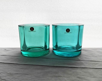 May include: Two turquoise-colored glass votive candle holders. Each has a cylindrical shape with a thick base. The text "MADE IN FINLAND" is printed on each holder. The candle holders are displayed on a gray surface.