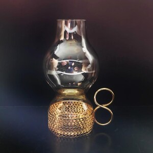 Puede incluir: Una l&aacute;mpara de hurac&aacute;n de cristal con una chimenea superior ahumada y una base transparente. La base est&aacute; envuelta en una malla met&aacute;lica dorada e incluye un asa decorativa de metal dorado con forma de ocho.