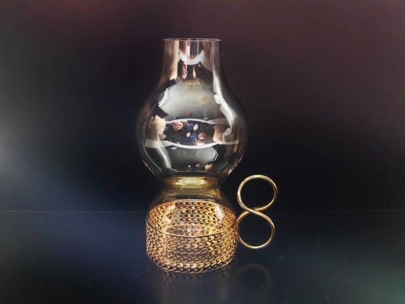 Puede incluir: Una l&aacute;mpara de hurac&aacute;n de cristal con una chimenea superior ahumada y una base transparente. La base est&aacute; envuelta en una malla met&aacute;lica dorada e incluye un asa decorativa de metal dorado con forma de ocho.
