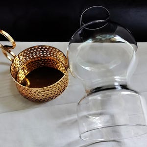 Puede incluir: Un portavasos de metal dorado con un patr&oacute;n de celos&iacute;a decorativo y un asa en forma de bucle, colocado junto a un inserto de vidrio transparente y redondeado dise&ntilde;ado para encajar en el soporte, con una base ancha y un cuello estrecho.