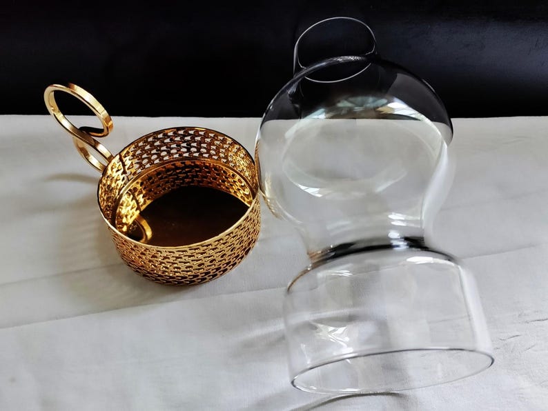 Puede incluir: Un portavasos de metal dorado con un patr&oacute;n de celos&iacute;a decorativo y un asa en forma de bucle, colocado junto a un inserto de vidrio transparente y redondeado dise&ntilde;ado para encajar en el soporte, con una base ancha y un cuello estrecho.