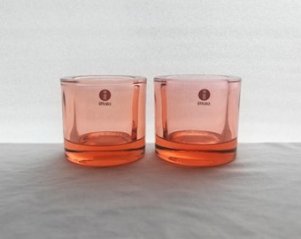 May include: Two cylindrical glass candle holders in a translucent salmon pink hue. Each piece features a thick, solid base and a smooth, clear glass wall. The Iittala logo, consisting of a red circle with a white letter i, is printed on the front.