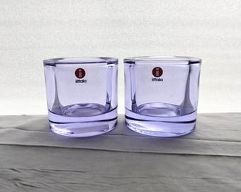 May include: Two clear, square-shaped glass candle holders with a light purple hue. Each holder has the "Iittala" logo in red. The candle holders are sitting on a light gray surface.