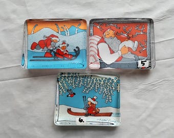 Puede incluir: Tres adornos de vidrio con escenas navideñas de invierno. El primer adorno presenta a dos personas con ropa roja y blanca montando un trineo tirado por un reno. El segundo adorno presenta a una persona con ropa roja y blanca sentada en un sofá con un libro. El tercer adorno presenta a una persona con ropa roja y blanca montando un trineo tirado por un reno. Los adornos están numerados 5 y 6.