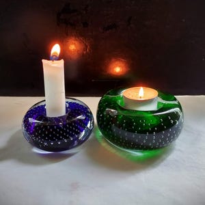 Op de afbeelding: Twee glazen kaarsenstandaards, een blauwe en een groene, met een witte kaars in de blauwe standaard en een theelicht in de groene standaard.