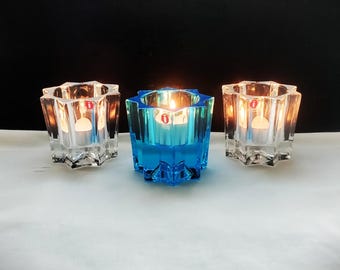 Puede incluir: Tres portavelas de cristal en forma de estrella dispuestos en fila. Dos portavelas son de cristal transparente y el central es de cristal azul vibrante. Cada pieza tiene una pequeña pegatina circular roja con una letra i blanca en el lateral.