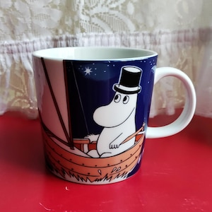May include: A white ceramic mug featuring a dark blue night sky background with a white Moomin character wearing a black top hat, sitting in a small wooden boat with a sail. The mug has a white interior and a white handle on the right side.