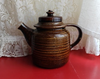 Puede incluir: Una tetera de cerámica marrón con tapa y asa. La tetera tiene un patrón de rayas horizontales texturizadas en tonos marrones y está colocada sobre una superficie roja. La tetera está diseñada para preparar té.