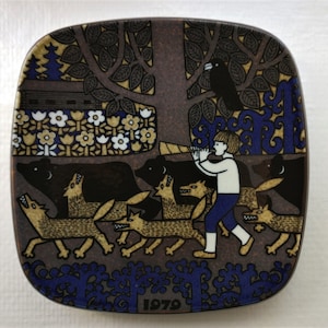 Puede incluir: Un plato cuadrado de cerámica con fondo marrón y una ilustración colorida de un niño tocando un cuerno en un bosque con animales. El plato tiene un borde azul con un diseño floral y el año 1979 impreso en el centro inferior.