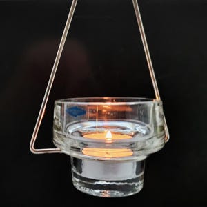 Puede incluir: Un portavelas de vidrio transparente con un marco de suspensión de metal. El portavelas tiene una base redonda y una parte superior cilíndrica. Una pequeña vela blanca está ardiendo dentro del portavelas.