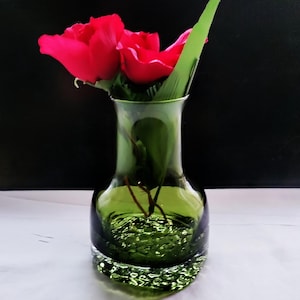 May include: A green glass vase with a textured base and a flared top, holding two vibrant red roses and green leaves. The vase has a dark green hue and sits on a white surface against a black background.