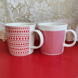 Può includere: Due tazze in ceramica bianca con disegni rossi. Una tazza ha un motivo a pois rossi e l'altra ha strisce rosse.
