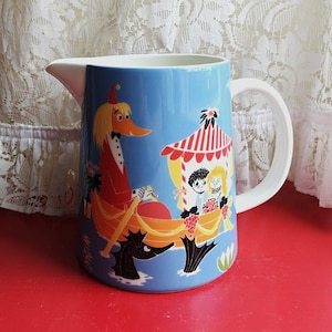 May include: A blue ceramic pitcher with a white handle and spout. The pitcher features a colorful illustration of a duck-like character in a boat with other characters. The design includes a red and white striped tent and other whimsical elements.
