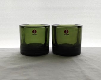 May include: Two cylindrical olive green glass tumblers with thick bases. Each glass features a small red and white circular sticker on the front with the letter i and the text iittala printed in black, indicating the brand of the glassware.