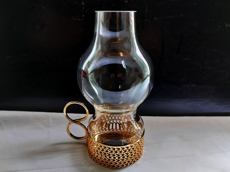 Puede incluir: Una pantalla de l&aacute;mpara de hurac&aacute;n de cristal transparente con una parte superior bulbosa y una base cil&iacute;ndrica. La base est&aacute; recubierta por una funda de metal dorado con un patr&oacute;n geom&eacute;trico recortado y un asa circular lateral.