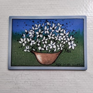 Op de afbeelding: Een rechthoekige keramische tegel met een bloemstilleven. Het kunstwerk toont een bruine vaas vol witte bloemen tegen een groene en blauwe achtergrond, omlijst door een lichtblauwe rand. De handtekening van de kunstenaar is zichtbaar.