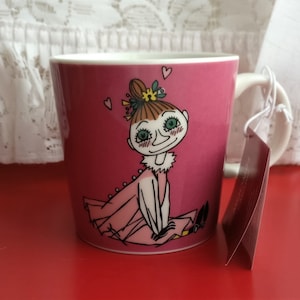 May include: A pink ceramic mug with a white handle features a cartoon illustration of a girl with brown hair, green eyes, and a pink dress. The girl is wearing a white collar and has a flower in her hair. The mug has a pink background with white hearts.