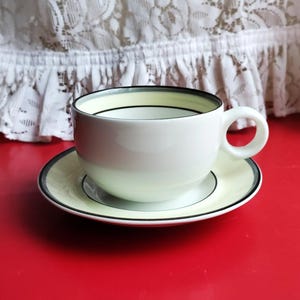May include: A white teacup and saucer with a black and yellow trim. The saucer has a yellow band with a black line on either side. The teacup has a yellow band with a black line on the top.