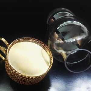 Puede incluir: Un portavasos de metal dorado con un exterior texturizado estilo tejido y un asa curva, junto con un inserto de vidrio transparente y redondeado. El soporte de metal presenta una superficie interior lisa y reflectante y un borde decorativo.