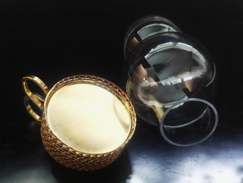 Puede incluir: Un portavasos de metal dorado con un exterior texturizado estilo tejido y un asa curva, junto con un inserto de vidrio transparente y redondeado. El soporte de metal presenta una superficie interior lisa y reflectante y un borde decorativo.