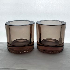 Op de afbeelding: Twee heldere glazen kaarshouders met een bruine tint. De kaarshouders zijn cilindrisch van vorm en hebben een licht uitlopende lip.