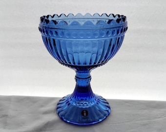 Op de afbeelding: Een blauwe glazen kom met een voet. De kom heeft een gegolfde rand en een getextureerd ontwerp. De voet heeft het woord "Iittala" erop gedrukt.