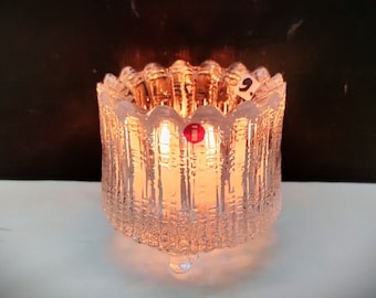 May include: A clear glass candle holder with a textured design and three legs. The candle holder is lit and the flame is visible.
