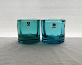 May include: Two teal-colored glass votive candle holders with thick, clear bases. Each piece features a small, circular red and white sticker on the front with the text iittala printed in black. The glass has a smooth, cylindrical shape.
