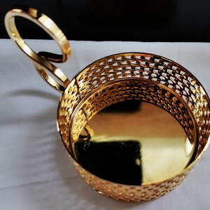 Puede incluir: Un portavasos de metal dorado con una base s&oacute;lida pulida y una pared lateral con un patr&oacute;n de celos&iacute;a decorativo. El soporte cuenta con un asa &uacute;nica en forma de bucle curvo que se extiende desde el lateral, dando una silueta distintiva.